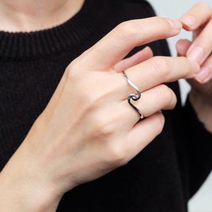 May include: A silver wave ring with a textured finish. The ring is worn on the index finger of a hand.