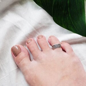 Puede incluir: Un anillo de dedo del pie de plata con un diseño decorativo. El anillo está en el dedo del pie de una persona con esmalte de uñas marrón claro.