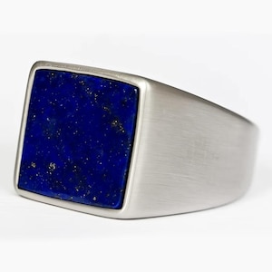 以下が含まれることがあります： 正方形の深い青色のラピスラズリの石が付いた、銀色の指輪。指輪はつや消し仕上げの金属で、太いバンドが付いています。ラピスラズリの石には金色の斑点があります。大胆なジュエリーです。