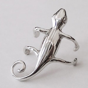 Puede incluir: Pendiente de oreja de camaleón de plata. El pendiente tiene forma de camaleón con la cola enroscada alrededor de la oreja. El camaleón está detallado con escamas texturizadas y tiene un aspecto realista.