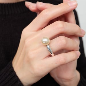 May include: A silver ring with a large, white pearl set in the center. The ring is worn on a finger.