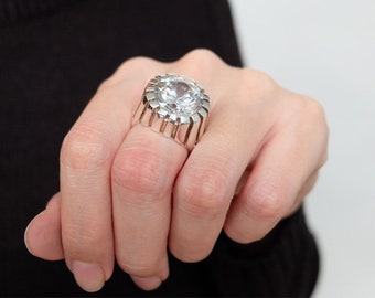 May include: A large, silver ring with a clear, faceted gemstone set in a circular bezel. The ring has a wide band with a textured, geometric design.