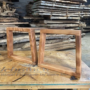 Puede incluir: Dos patas de mesa rectangulares de madera sin terminar, con un acabado de veta de madera natural. Las patas están anguladas y parecen hechas a mano. El fondo presenta pilas de tablas de madera en bruto, lo que sugiere un taller de carpintería.