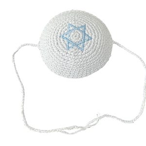 Può includere: Una kippah bianca all'uncinetto con una stella di David blu chiaro ricamata. La kippah ha una forma rotonda e un cordino bianco per fissarla. Lo sfondo è bianco. Copricapo religioso ebraico.
