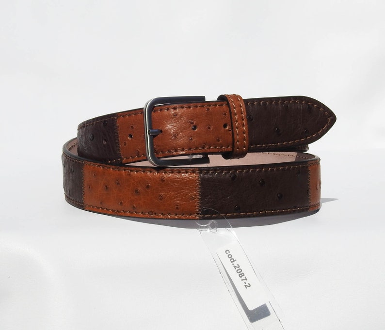 May include: A brown and black leather belt with an ostrich skin pattern. The belt has a silver buckle and is stitched with brown thread. The tag attached to the belt reads "cod.2087-2".
