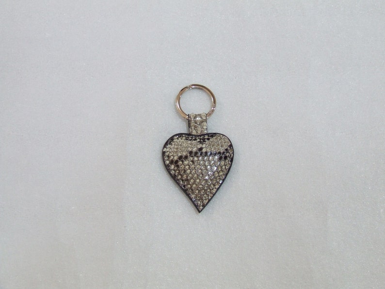 Op de afbeelding: Een zilveren sleutelhanger met een hartvormige hanger. De hanger is bedekt met strass steentjes en heeft een slangenhuidpatroon.