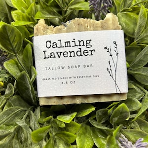Pu&ograve; includere: Una saponetta al sego rettangolare con l'etichetta "Calming Lavender" con un'etichetta bianca, immersa in un fogliame verde. L'etichetta include il testo "TALLOW SOAP BAR" e "3.5 OZ". La saponetta &egrave; di colore marrone chiaro ed &egrave; circondata da foglie verdi e fiori viola.
