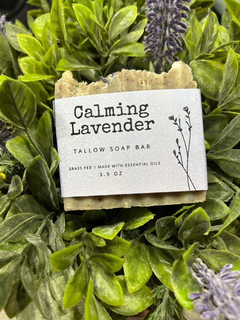 Pu&ograve; includere: Una saponetta al sego rettangolare con l'etichetta "Calming Lavender" con un'etichetta bianca, immersa in un fogliame verde. L'etichetta include il testo "TALLOW SOAP BAR" e "3.5 OZ". La saponetta &egrave; di colore marrone chiaro ed &egrave; circondata da foglie verdi e fiori viola.