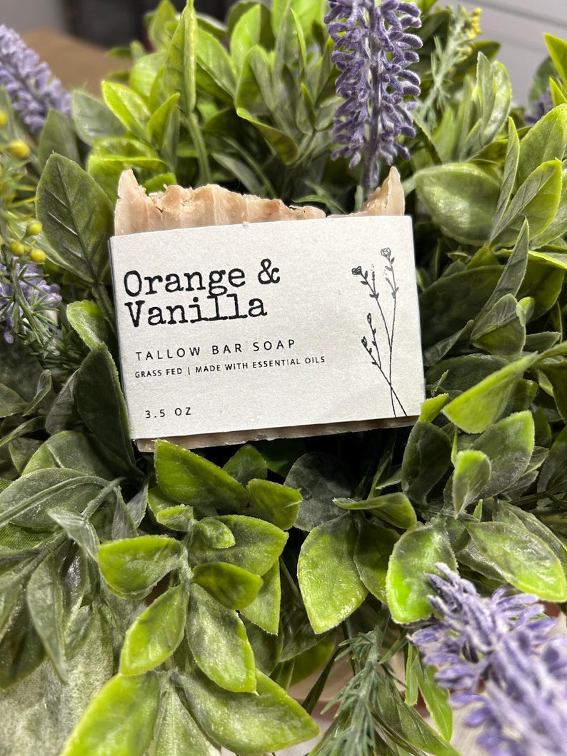 Pu&ograve; includere: Una saponetta al sego con la scritta "Orange & Vanilla" su un'etichetta bianca. L'etichetta include anche il testo "TALLOW BAR SOAP" e "GRASS FED | MADE WITH ESSENTIAL OILS". Il sapone pesa 99 grammi. Il sapone &egrave; circondato da foglie verdi e fiori viola.