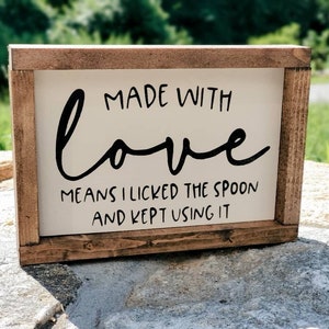 May include: A rectangular wooden sign with a white background and a brown frame. The sign features black cursive text that reads "MADE WITH love MEANS I LICKED THE SPOON AND KEPT USING IT."