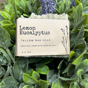 Pu&ograve; includere: Una saponetta al sego di limone ed eucalipto, con la scritta "Lemon Eucalyptus Tallow Bar Soap" su un'etichetta beige. L'etichetta indica anche "Grass Fed | Made with Essential Oils" e "3.5 OZ". Il sapone &egrave; circondato da fogliame verde e fiori viola.