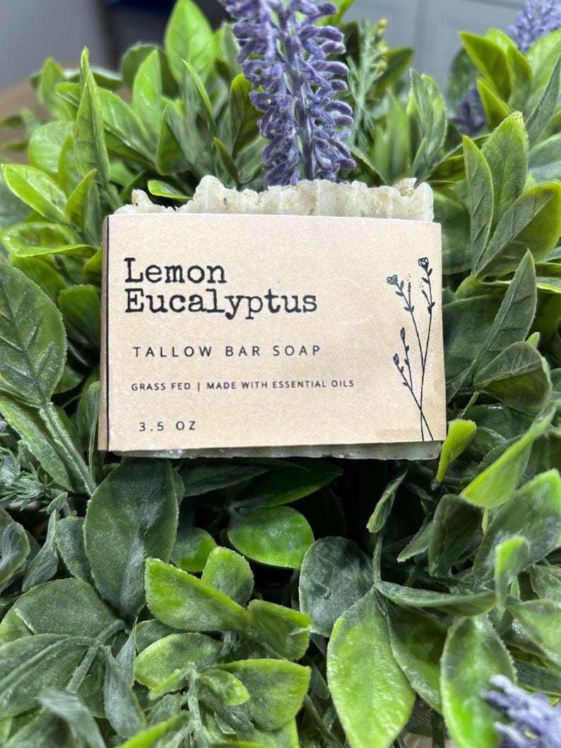 Pu&ograve; includere: Una saponetta al sego di limone ed eucalipto, con la scritta "Lemon Eucalyptus Tallow Bar Soap" su un'etichetta beige. L'etichetta indica anche "Grass Fed | Made with Essential Oils" e "3.5 OZ". Il sapone &egrave; circondato da fogliame verde e fiori viola.