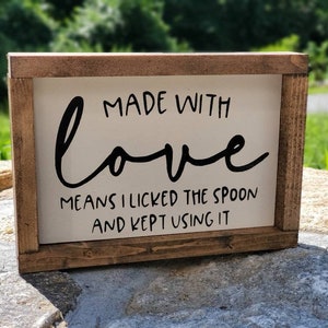May include: A rectangular wooden sign with a white background and a dark brown frame. The sign features the phrase "MADE WITH love MEANS I LICKED THE SPOON AND KEPT USING IT" in black lettering. The sign is displayed outdoors.