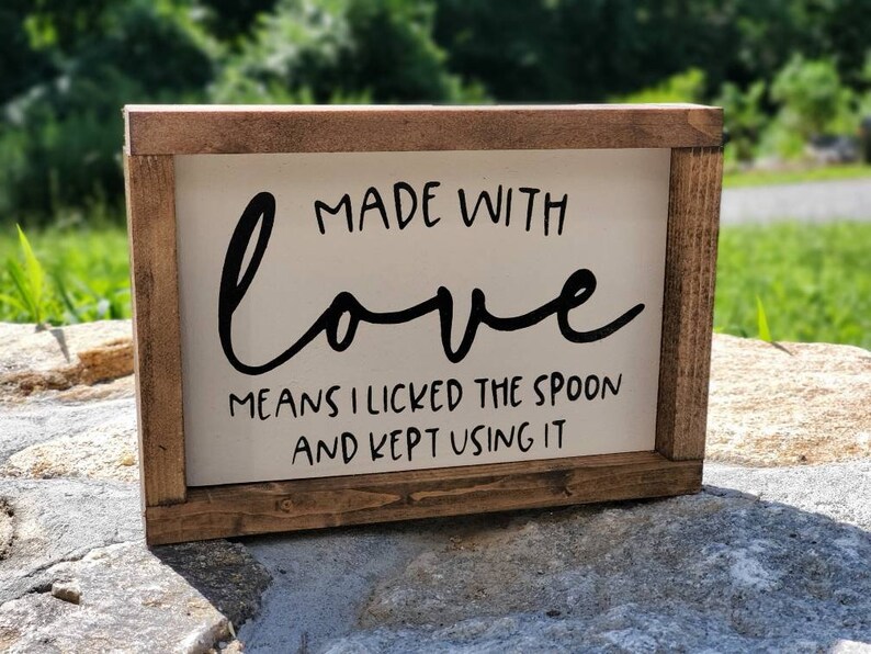 May include: A rectangular wooden sign with a white background and a dark brown frame. The sign features the phrase "MADE WITH love MEANS I LICKED THE SPOON AND KEPT USING IT" in black lettering. The sign is displayed outdoors.