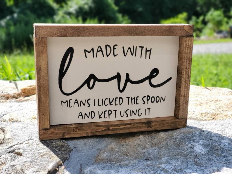 May include: A rectangular wooden sign with a white background and a brown frame. The sign features black cursive text that reads "MADE WITH love MEANS I LICKED THE SPOON AND KEPT USING IT."