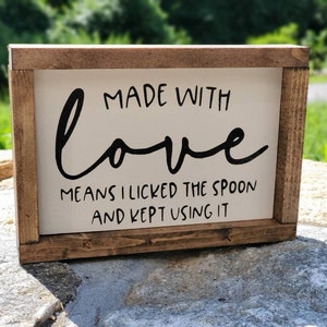 May include: A rectangular wooden sign with a white background and a brown frame. The sign features black cursive text that reads "MADE WITH love MEANS I LICKED THE SPOON AND KEPT USING IT."