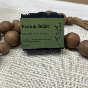 Pu&ograve; includere: Una saponetta scura con la scritta "Pine & Cedar" su un'etichetta verde. L'etichetta recita anche "TALLOW BAR SOAP" e "GRASS FED | MADE WITH ESSENTIAL OILS AND ACTIVATED CHARCOAL". Il sapone pesa circa 99 grammi.