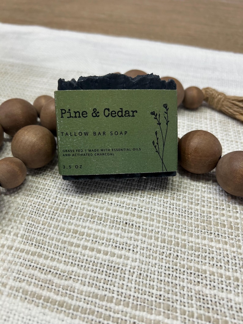 Pu&ograve; includere: Una saponetta scura con la scritta "Pine & Cedar" su un'etichetta verde. L'etichetta recita anche "TALLOW BAR SOAP" e "GRASS FED | MADE WITH ESSENTIAL OILS AND ACTIVATED CHARCOAL". Il sapone pesa circa 99 grammi.