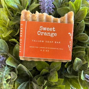 Pu&ograve; includere: Una saponetta al sego rettangolare con una superficie strutturata e un'etichetta arancione che recita "Sweet Orange". L'etichetta indica anche "TALLOW SOAP BAR" e "4.5 OZ". Il sapone &egrave; circondato da foglie verdi e viola.