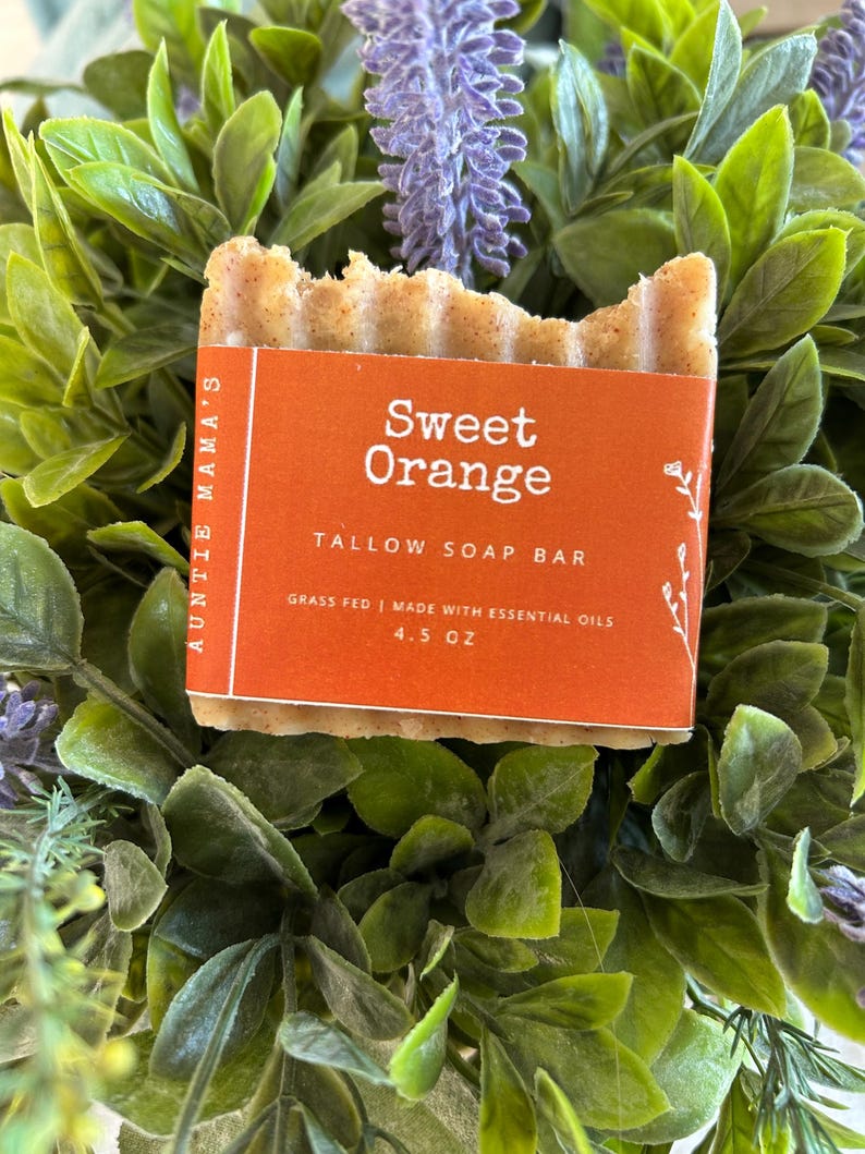 Pu&ograve; includere: Una saponetta al sego rettangolare con una superficie strutturata e un'etichetta arancione che recita "Sweet Orange". L'etichetta indica anche "TALLOW SOAP BAR" e "4.5 OZ". Il sapone &egrave; circondato da foglie verdi e viola.