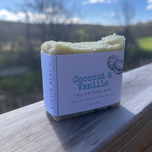 Pu&ograve; includere: Una saponetta di sego di colore chiaro con un'etichetta che recita "Coconut & Vanilla". L'etichetta indica anche "Grass Fed | Made with Essential Oils" e "4.5 OZ". Il sapone &egrave; su una superficie di legno con uno sfondo sfocato.