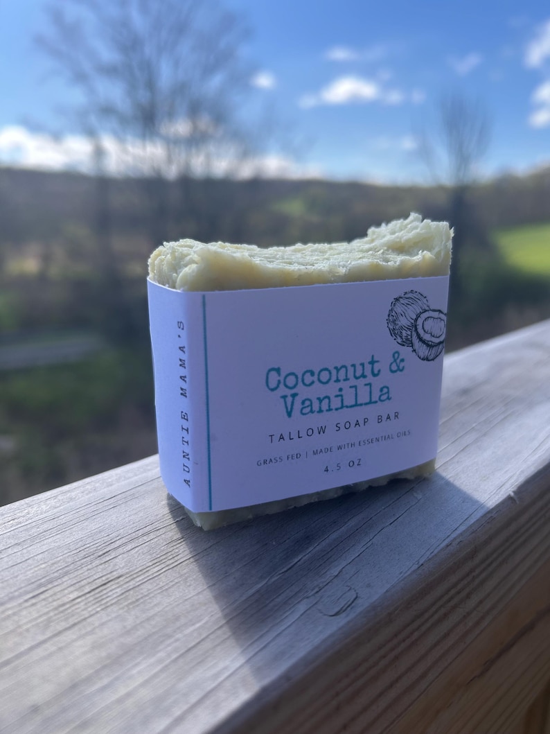 Pu&ograve; includere: Una saponetta di sego di colore chiaro con un'etichetta che recita "Coconut & Vanilla". L'etichetta indica anche "Grass Fed | Made with Essential Oils" e "4.5 OZ". Il sapone &egrave; su una superficie di legno con uno sfondo sfocato.