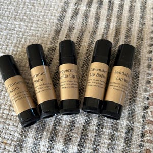Pu&ograve; includere: Cinque tubetti di balsamo per le labbra con tappi neri ed etichette color beige sono disposti su una superficie strutturata. Le etichette recitano: Black Cinnamon Balm, Peppermint Balm, Peppermint Vanilla Lip, Lavender Lip Balm e Sandalwood Lip Balm.