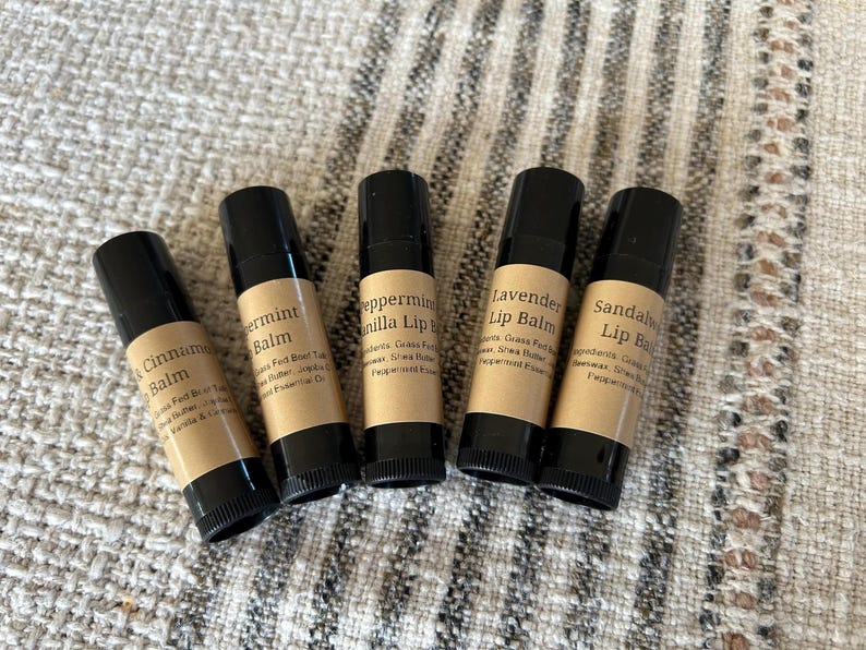 Pu&ograve; includere: Cinque tubetti di balsamo per le labbra con tappi neri ed etichette color beige sono disposti su una superficie strutturata. Le etichette recitano: Black Cinnamon Balm, Peppermint Balm, Peppermint Vanilla Lip, Lavender Lip Balm e Sandalwood Lip Balm.