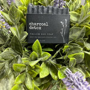 Pu&ograve; includere: Un sapone in saponetta al carbone detox grigio scuro con un'etichetta nera. L'etichetta recita "charcoal detox TALLOW BAR SOAP GRASS FED | MADE WITH ESSENTIAL OILS 3.5 OZ". Il sapone &egrave; circondato da foglie verdi e fiori viola.