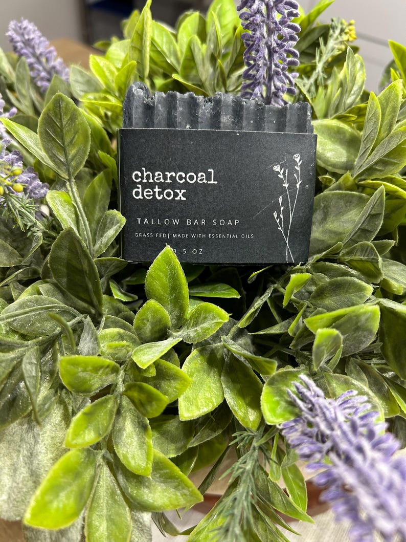 Pu&ograve; includere: Un sapone in saponetta al carbone detox grigio scuro con un'etichetta nera. L'etichetta recita "charcoal detox TALLOW BAR SOAP GRASS FED | MADE WITH ESSENTIAL OILS 3.5 OZ". Il sapone &egrave; circondato da foglie verdi e fiori viola.