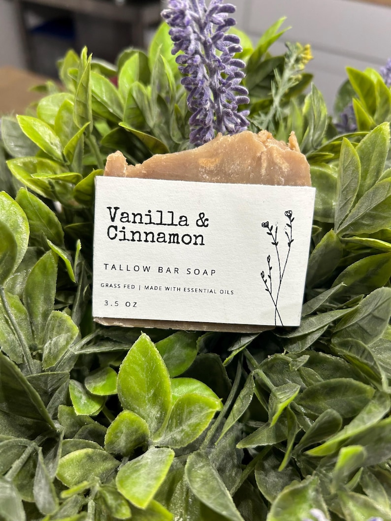 Pu&ograve; includere: Un sapone in barra al sego marrone chiaro con un'etichetta bianca che recita "Vanilla & Cinnamon". L'etichetta indica anche "Grass Fed | Made with Essential Oils" e "3.5 OZ". Il sapone &egrave; circondato da foglie verdi e fiori viola.
