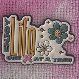 Könnte beinhalten: Ein weißer, gelber und türkisfarbener Emaille-Pin mit den Worten "ENJOY LIFE ONE AT A TIME!" und Blumengrafiken. Der Pin ist vor einem rosa-lila Batik-Hintergrund platziert.
