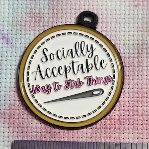 May include: A gold-colored metal embroidery hoop with a white background and black stitching around the edge. The hoop has a pink and black inscription that reads "Socially Acceptable Way to Stab Things" with a needle and thread image in the center.