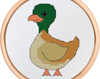 Puede incluir: Un diseño de punto de cruz de un pato en un bastidor de bordado de madera. El pato tiene una cabeza verde, pico y patas naranjas, y un cuerpo en tonos beige y marrón. El fondo es blanco.