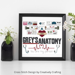 Może przedstawiać: Oprawione dzieło sztuki haftu krzyżykowego z napisem "Grey's Anatomy" dużymi literami. Projekt zawiera symbole medyczne, serca i frazy z serialu. Rama jest czarna, a tło białe.