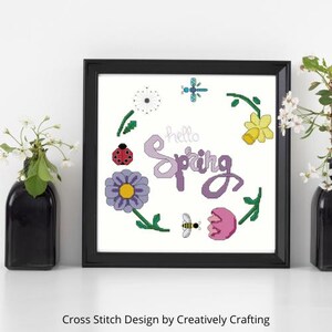 Puede incluir: Un diseño de punto de cruz enmarcado en un marco negro. El diseño presenta las palabras "hello Spring" en morado, rodeadas de flores coloridas, una mariquita, una abeja y una libélula. El diseño está sobre un fondo blanco.