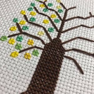 May include: Close-up of a brown and green cross-stitch embroidery of a tree with a thick trunk and branches with yellow and green leaves.