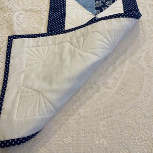 May include: A quilted table runner featuring a white base and a central diamond design in various shades of blue. The diamond is framed by a navy blue border with white polka dots. The underside is white with a fan-shaped quilted pattern.