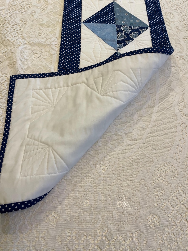 May include: A quilted table runner featuring a white base and a central diamond design in various shades of blue. The diamond is framed by a navy blue border with white polka dots. The underside is white with a fan-shaped quilted pattern.