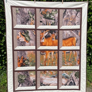 May include: A quilted blanket featuring a deer scene framed by a windowpane design. The central image showcases two deer in a forest setting, surrounded by a camouflage pattern and brown borders. The quilt has a cream-colored border.