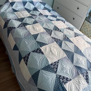 May include: A blue and white quilt with a swirling pattern. The quilt is made of various shades of blue and white fabrics and is quilted with a decorative stitch.