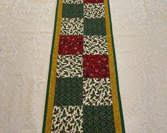 May include: A rectangular Christmas table runner with a patchwork design. The runner features alternating squares of red, green, and white fabric with holly and geometric patterns. It is bordered by a gold and green trim.