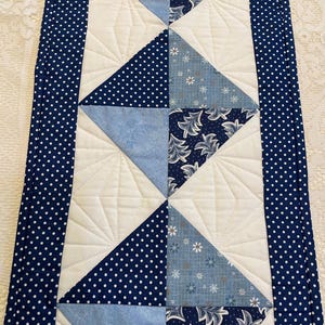 May include: A quilted table runner with a central design of alternating blue triangles. Some triangles are solid, others have white polka dots, and some feature floral patterns. The runner is bordered with a navy blue fabric with white polka dots.