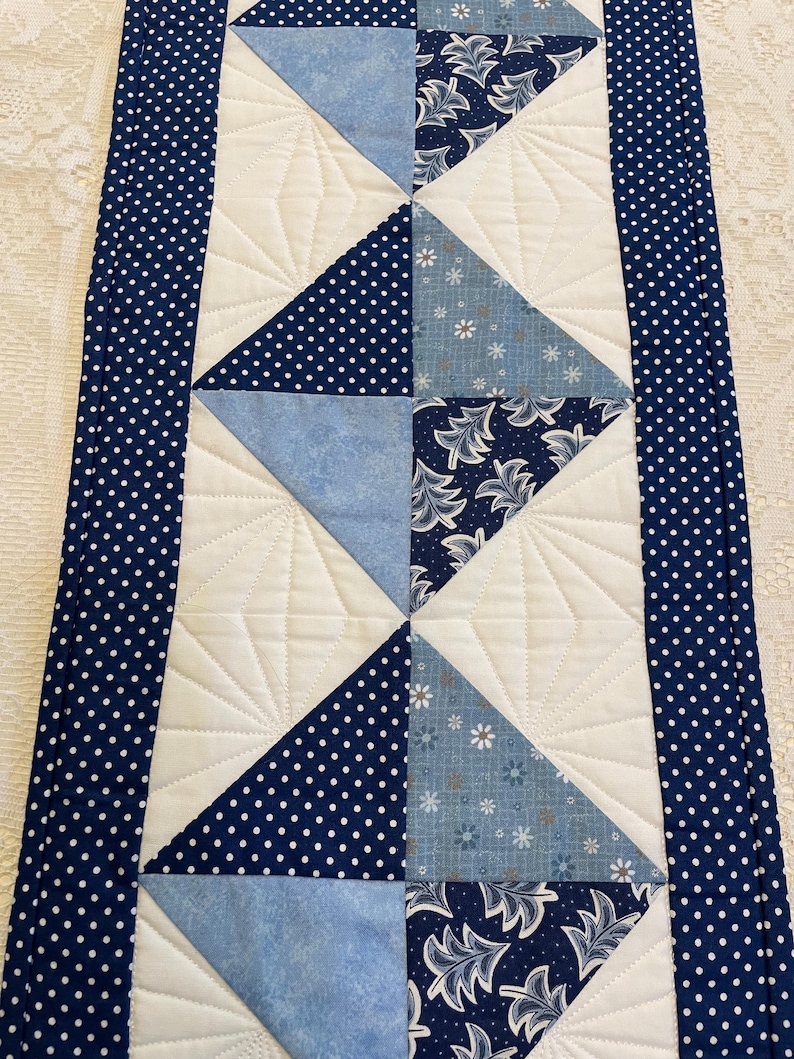 May include: A quilted table runner with a central design of alternating blue triangles. Some triangles are solid, others have white polka dots, and some feature floral patterns. The runner is bordered with a navy blue fabric with white polka dots.