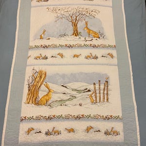 May include: A white and blue quilt with a winter scene featuring brown rabbits and a bare tree. The quilt has a white border and a blue backing.