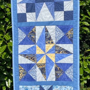 May include: A blue and white quilted table runner with star patterns. The runner features various shades of blue fabric, including solid and patterned designs. The stars are arranged in three sections, with a light blue border.