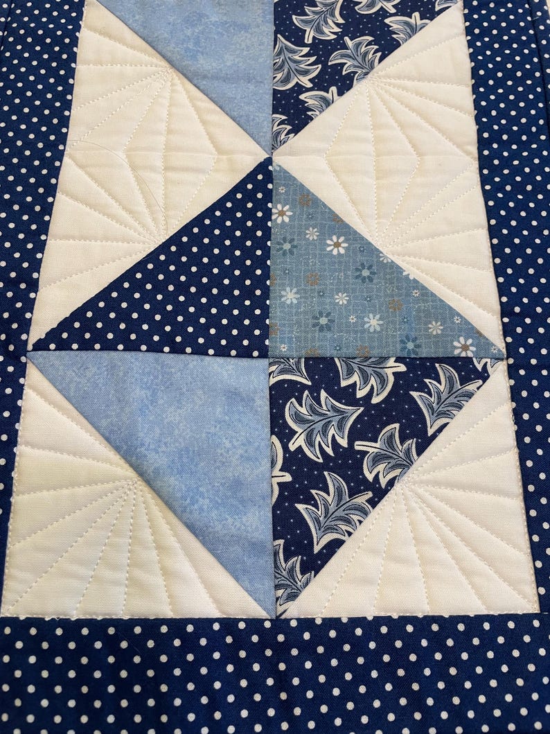 May include: A quilted table runner with a patchwork design. It features blue fabrics, including polka dots, floral patterns, and solid shades. White sections with stitched fan patterns provide contrast. The border is dark blue with white polka dots.