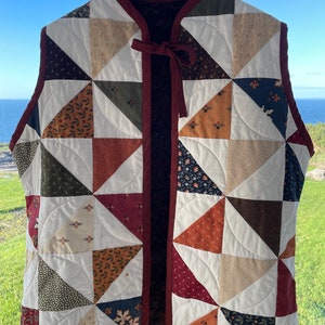 May include: A quilted vest with a patchwork design featuring various shades of brown, green, red, and blue. The vest has a tie closure at the neck and a burgundy trim.
