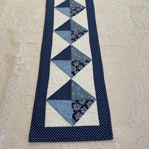 May include: A rectangular quilted table runner with a navy blue polka dot border. The center features alternating white and blue diamond shapes, with various blue patterned fabrics. The runner is likely handmade and adds a decorative touch to a table.