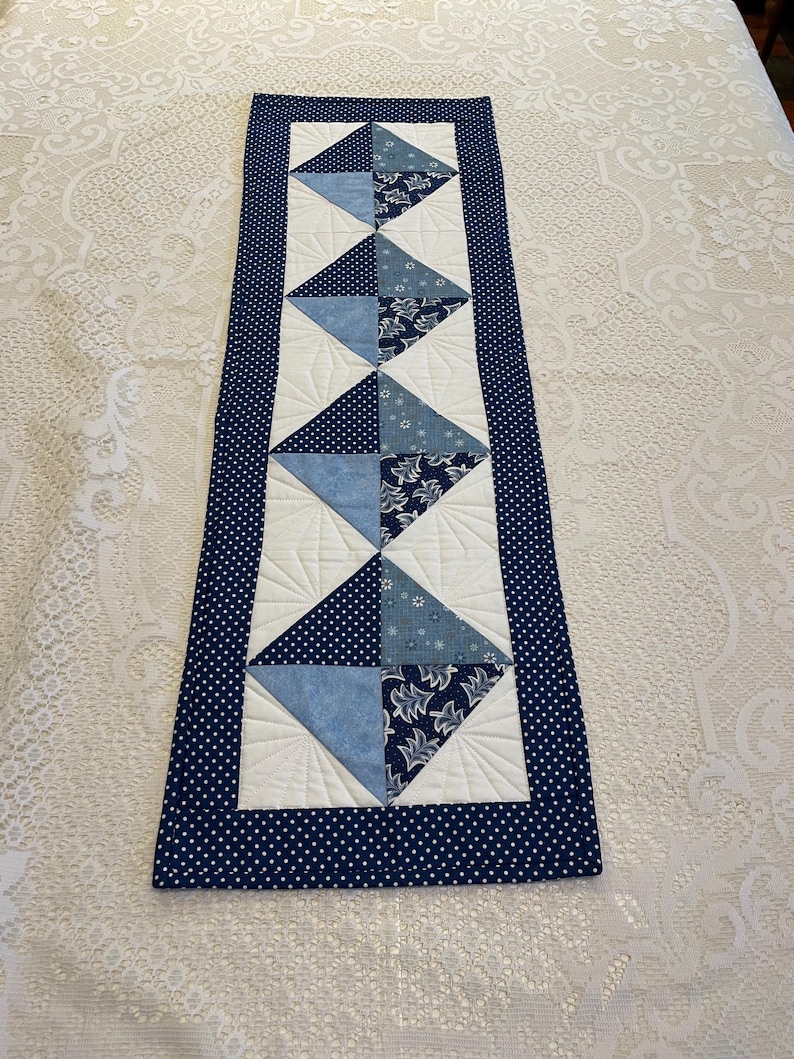 May include: A rectangular quilted table runner with a navy blue polka dot border. The centre features alternating white and blue diamond shapes, with various blue patterned fabrics. The runner is likely handmade and adds a decorative touch to a table.